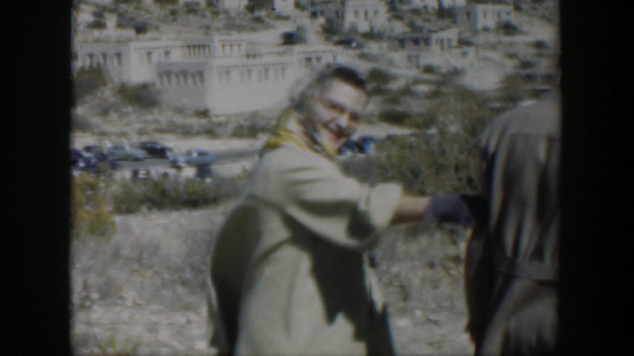 Smiling woman in a light coat gestures beside a man at the edge of a dry roadside landscape.