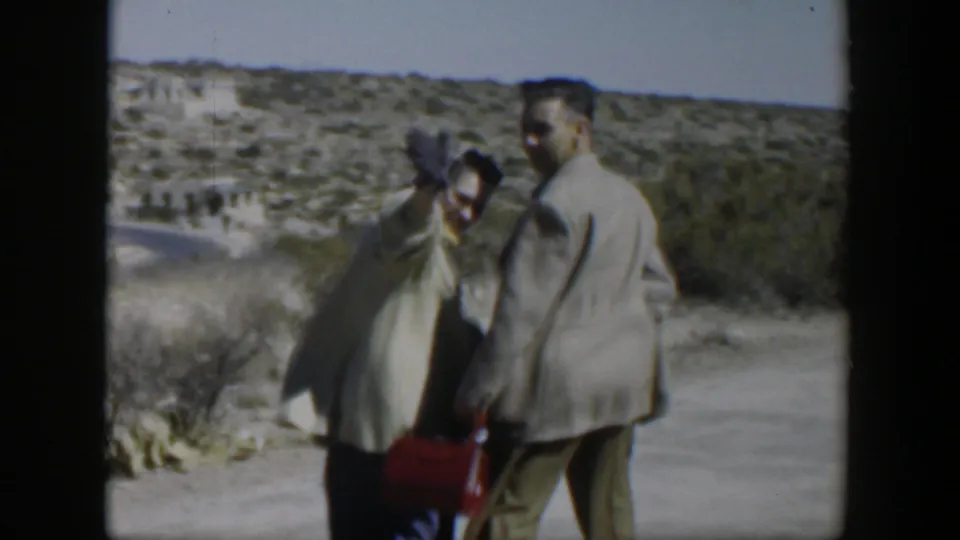 A couple stands close on a Texas roadside in 1951, with the woman lifting a hand or scarf near her face in bright light.