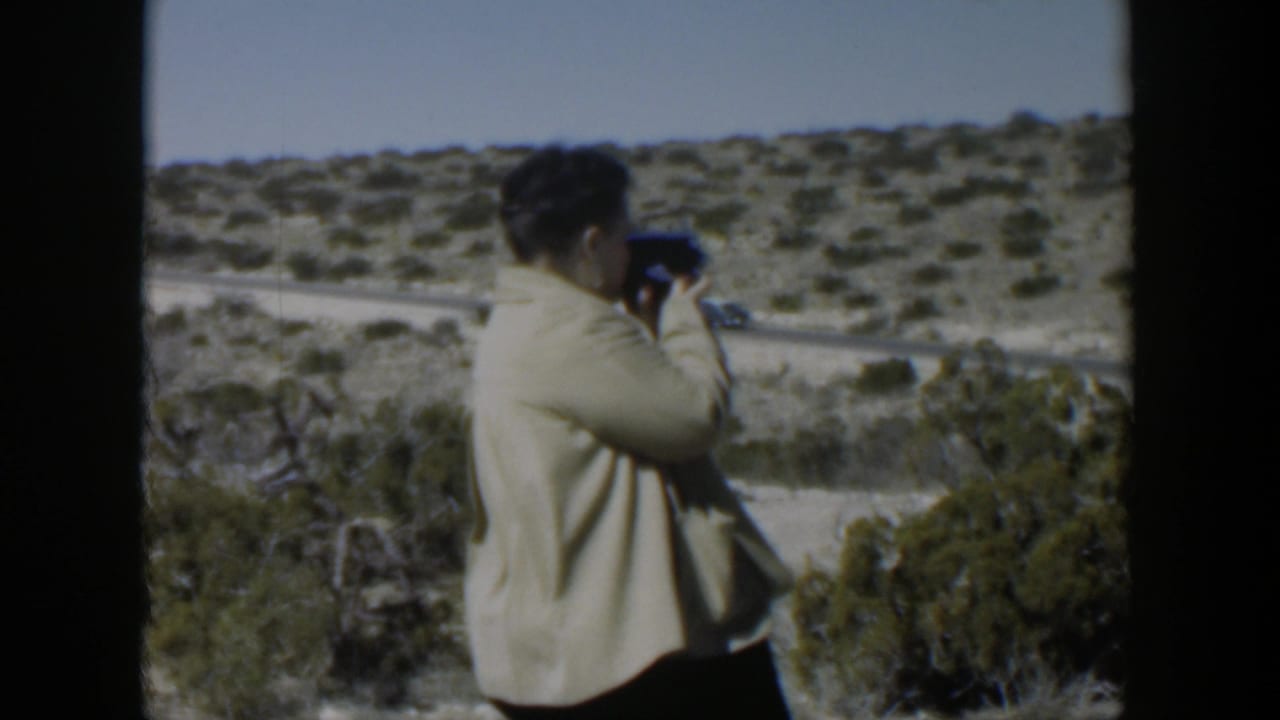 Woman in a light jacket films a scrubby hillside beside a dirt road.