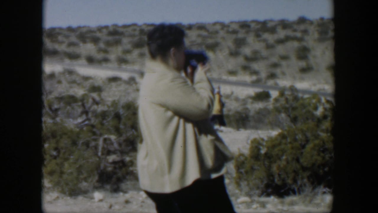 Woman holds a small camera toward a brushy desert road scene.