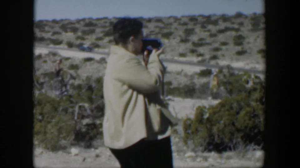 A woman continues filming from the hillside while a car moves along the dirt road in the 1951 Texas landscape behind her.
