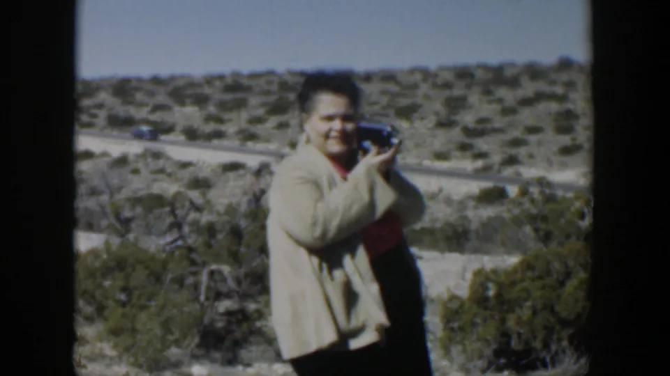 The woman turns toward the home-movie camera, smiling while holding her own small camera against a Texas desert roadside backdrop.
