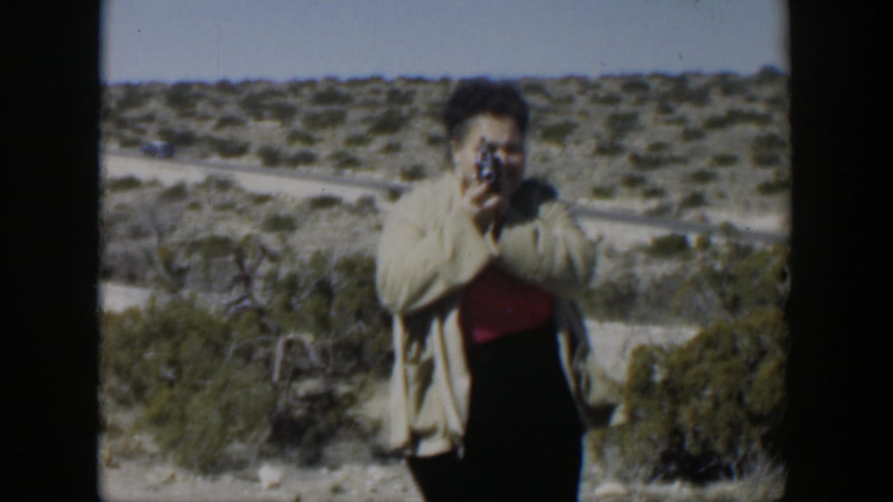 Woman in a light coat points a small camera toward the filmmaker in a dry landscape.