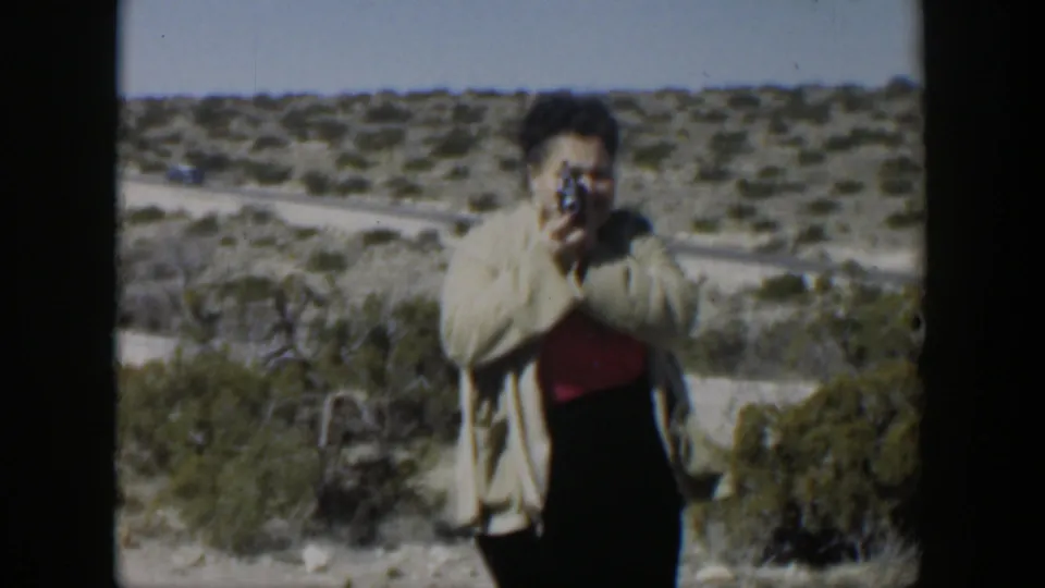 In metadata-supported 1951 Texas footage, a woman in a light coat aims a small camera across a scrubby desert overlook.