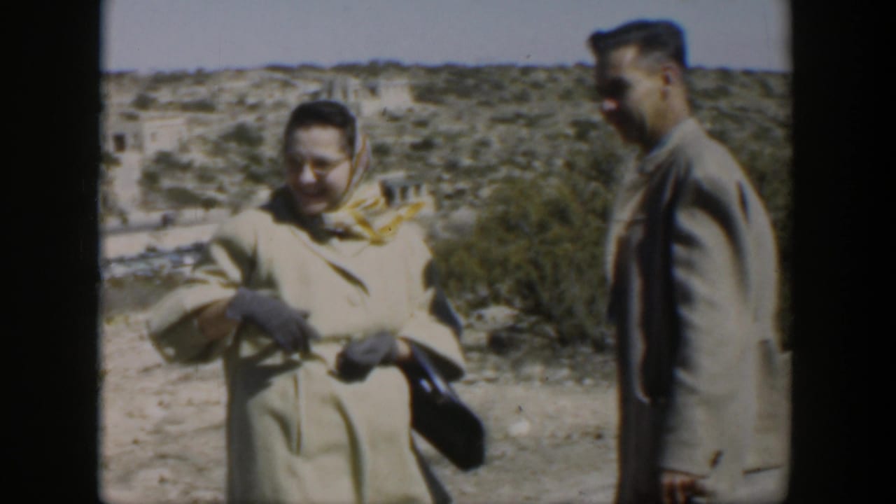 Woman in a headscarf and light coat stands beside a man on a desert path.