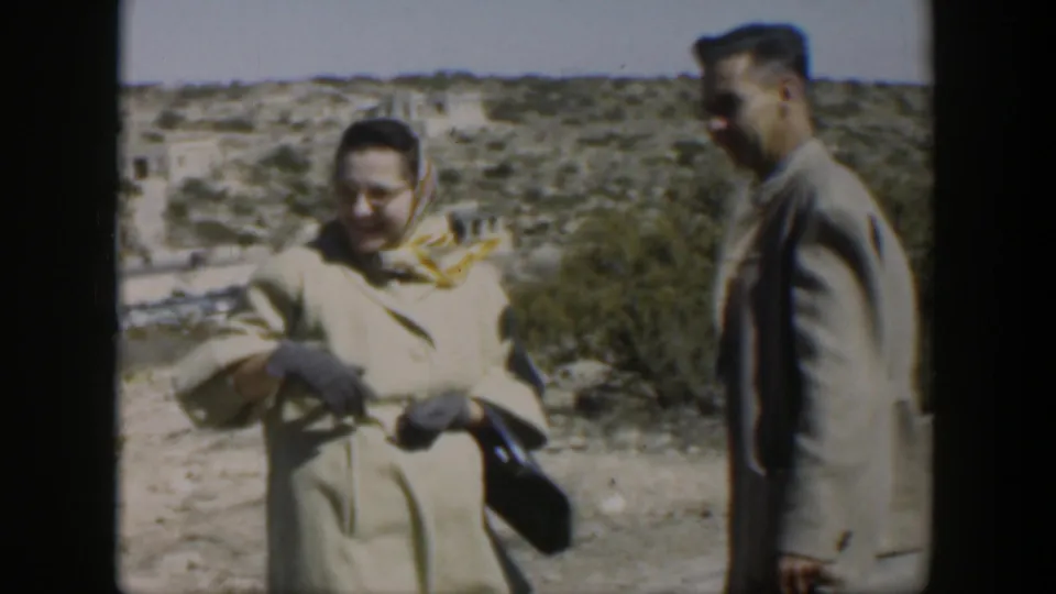 A woman in a headscarf and beige coat stands with a man in a light jacket in the 1951 Texas desert setting.
