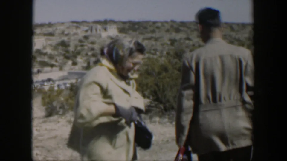 The woman and a man move along a dry path, with scrub vegetation and low distant structures behind them.