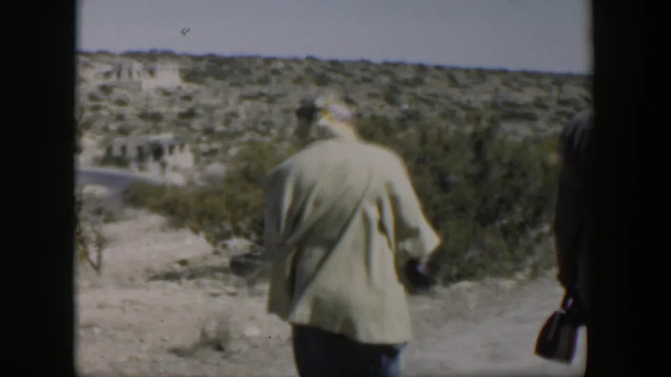 A man in a light jacket turns away on a desert path while another figure is partly visible at the frame edge.