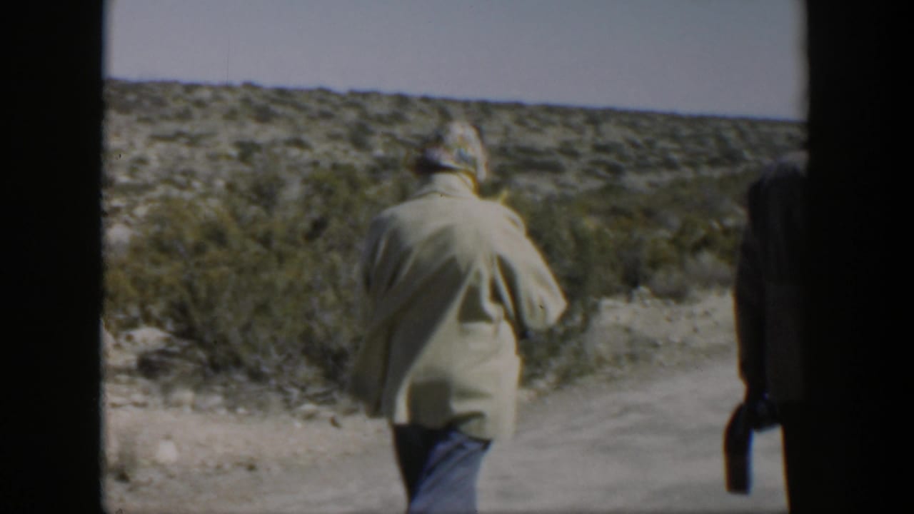 Two adults walk away along a dusty roadside with shrubs and open sky.