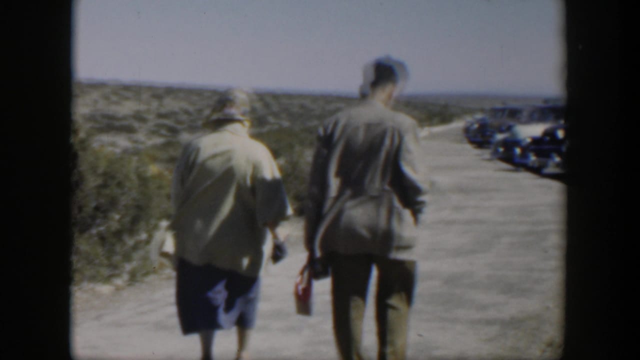 A man and woman walk toward parked classic cars on a roadside.