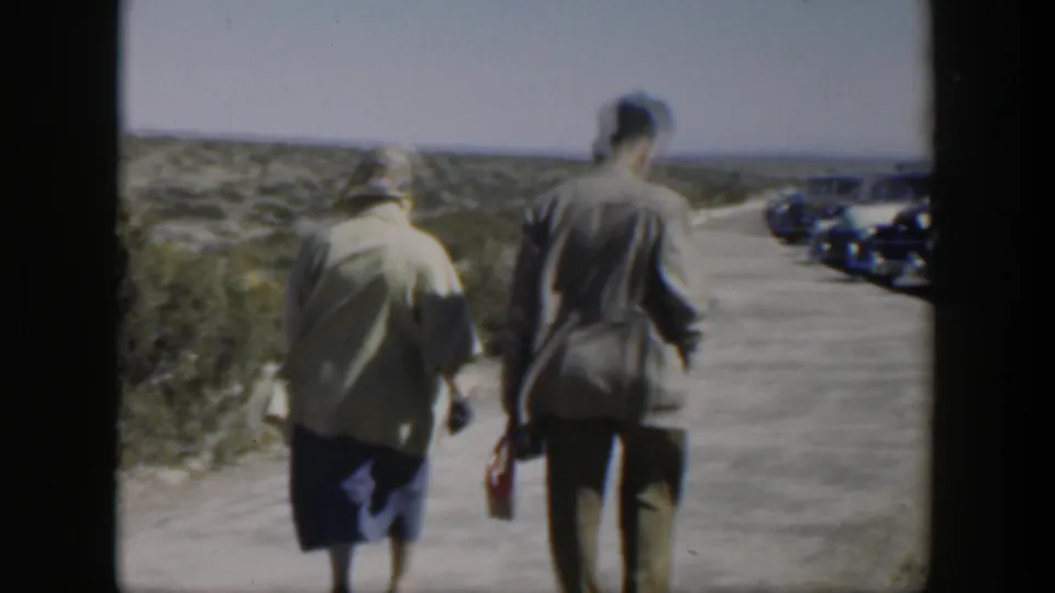 The man and woman continue along a Texas roadside toward a row of parked 1950s cars under clear daylight.