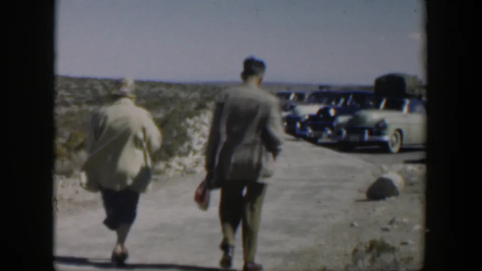 Two adults are seen from behind on a dusty Texas road, with several mid-century cars parked ahead beside low brush.