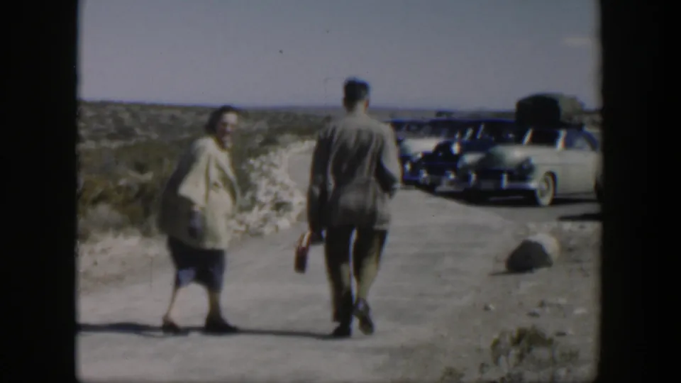 In the final frame, the woman turns back toward the camera while the man continues along the Texas roadside toward parked cars.