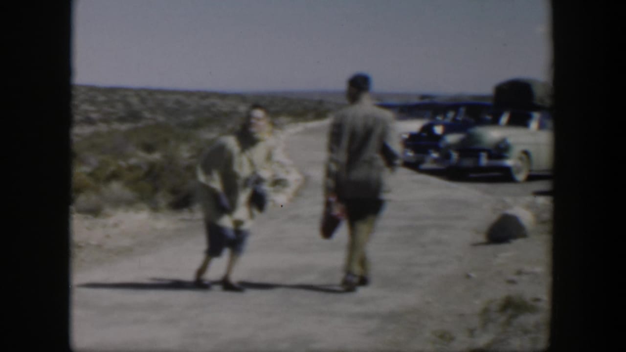 1951 Texas home-movie frame of a couple walking on a dusty road with parked mid-century cars nearby.