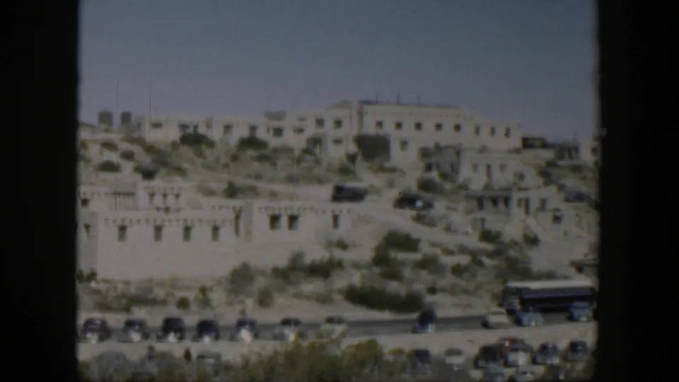 1951 Texas home-movie frame of hillside buildings with vintage vehicles and a larger vehicle near the road.