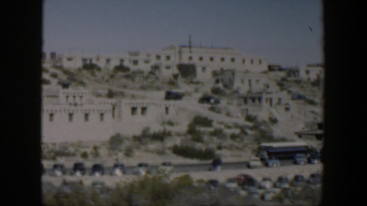 1951 Texas home-movie view of pale hillside buildings above a road with mid-century cars, a bus, shrubs, and bright sky.
