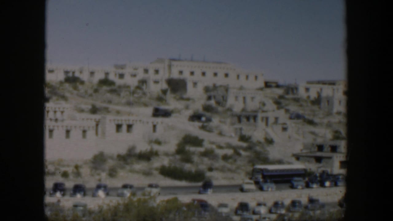 Distant Texas hillside scene with white adobe-style buildings, sparse greenery, road traffic, and a large bus in soft 8mm color.
