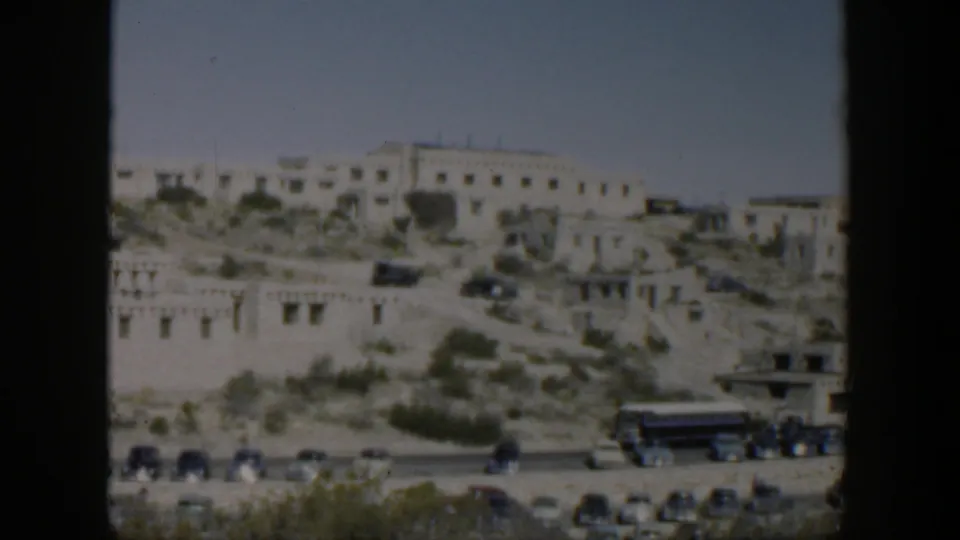 Distant Texas hillside scene with white adobe-style buildings, sparse greenery, road traffic, and a large bus in soft 8mm color.