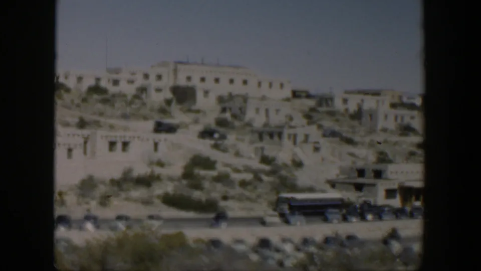 Soft 8mm frame of white buildings on a dusty Texas hillside, with a road, bus, cars, brush, and open sky visible.