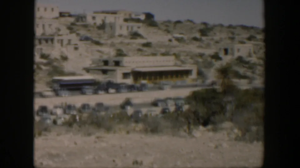 Soft 8mm frame of Texas hillside ruins with vintage cars and a low building near the road.