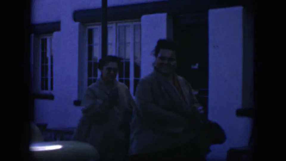 Blue-tinted nighttime 1951 home-movie frame of two women in coats smiling outside a building.