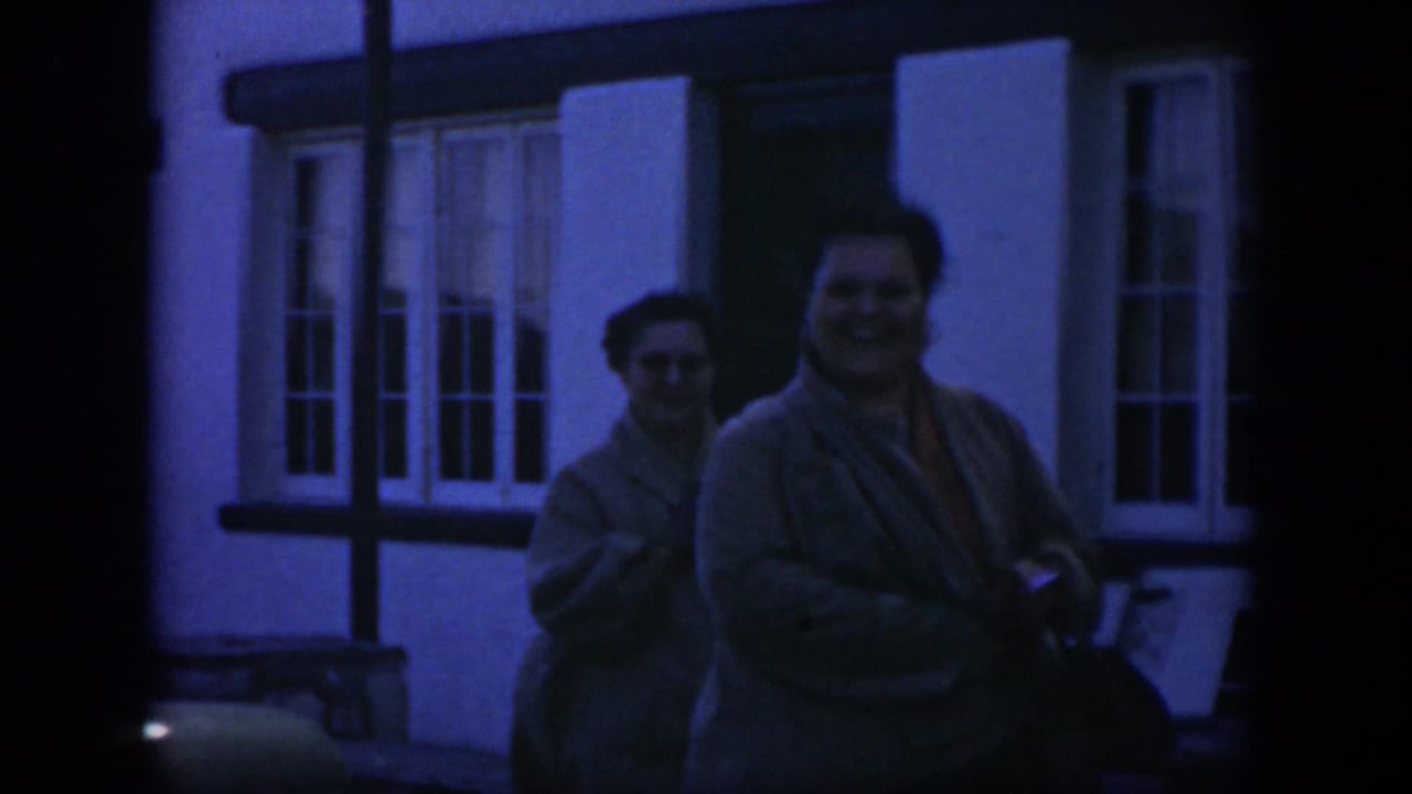 1951 Texas home-movie frame of two adult women in tan sweaters walking near a white building, with one woman smiling toward the camera.