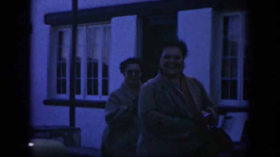 1951 Texas home-movie frame of two adult women in tan sweaters walking near a white building, with one woman smiling toward the camera.