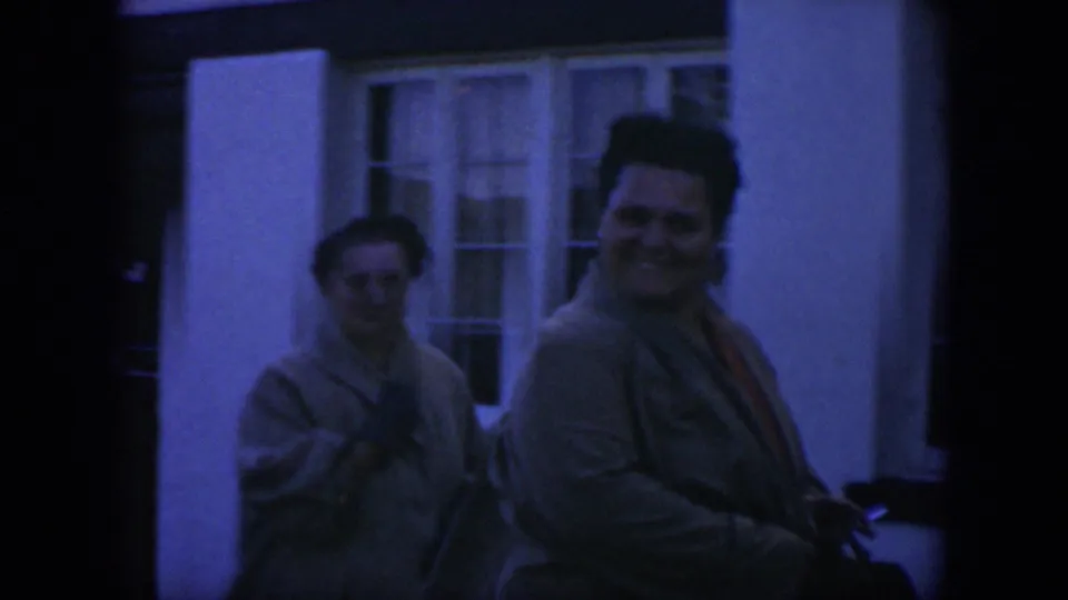 1951 Texas 8mm frame showing two adult women in sweaters walking outdoors beside a white building with white-framed windows.