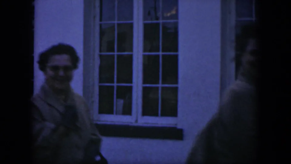 1951 Texas home-movie frame with one adult woman near a white-framed window and another person partly obscured at the edge.