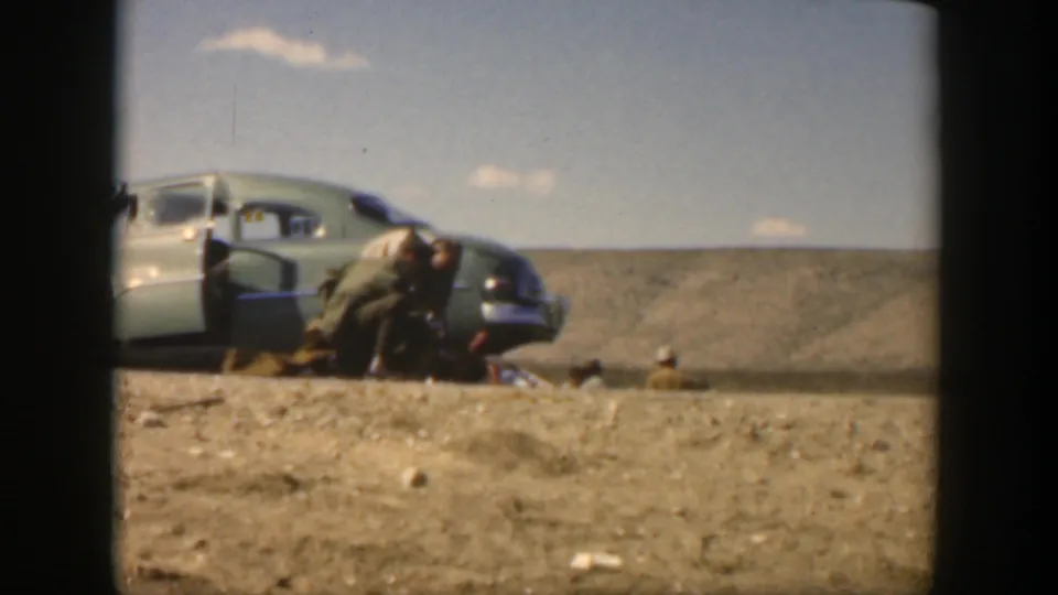 A brief adjacent 8mm frame shows a mid-century car with an open door on dry ground, with people crouched near the roadside.