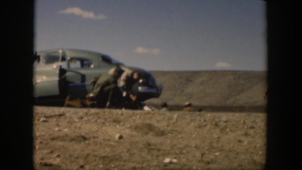1951 Texas home-movie frame showing soldiers giving roadside aid beside a light-colored sedan in a dry open landscape.