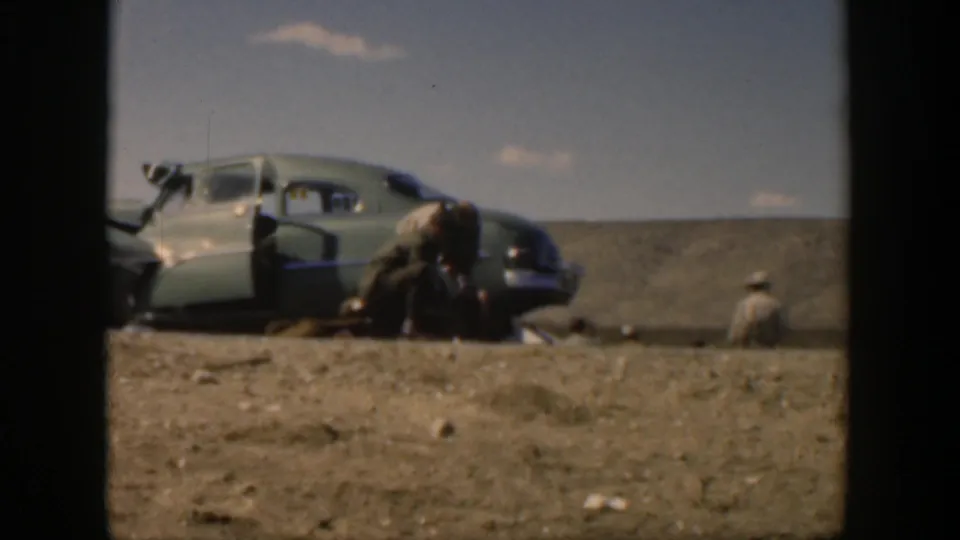 1951 Texas roadside accident frame with a soldier leaning toward an injured person beside a mid-century sedan.