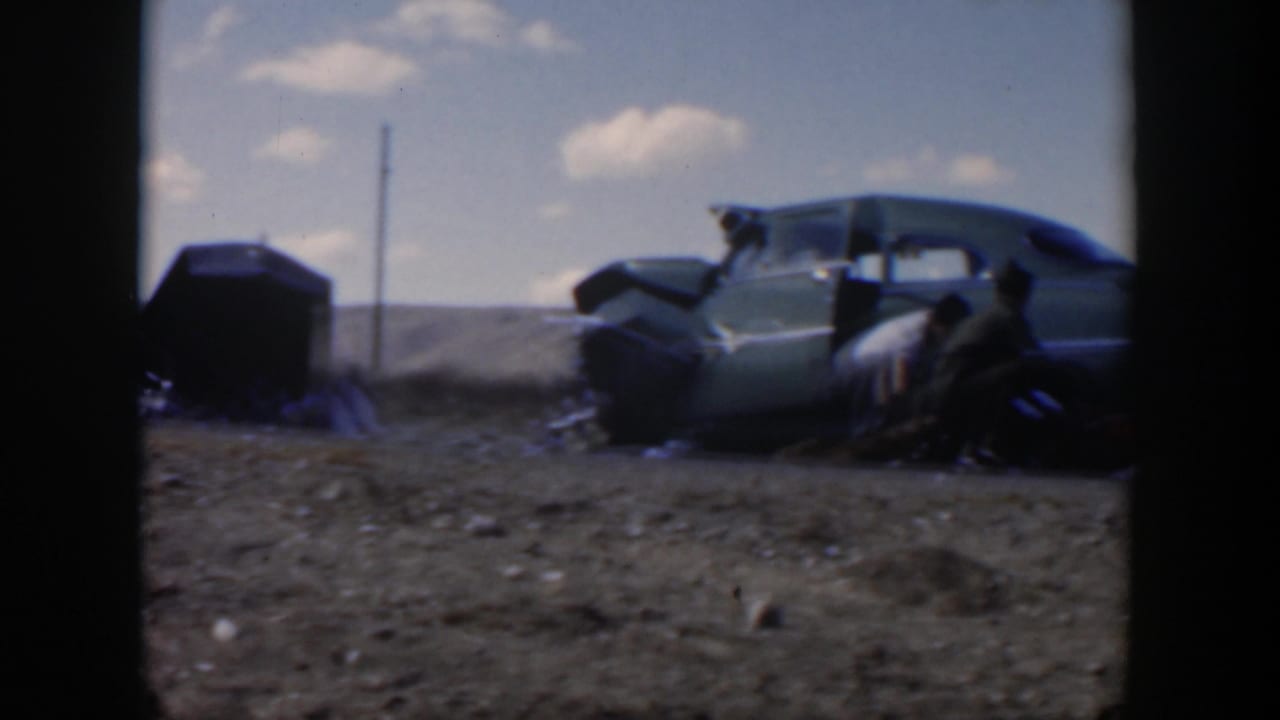 A green sedan sits by the road with men gathered near its side and rear area.