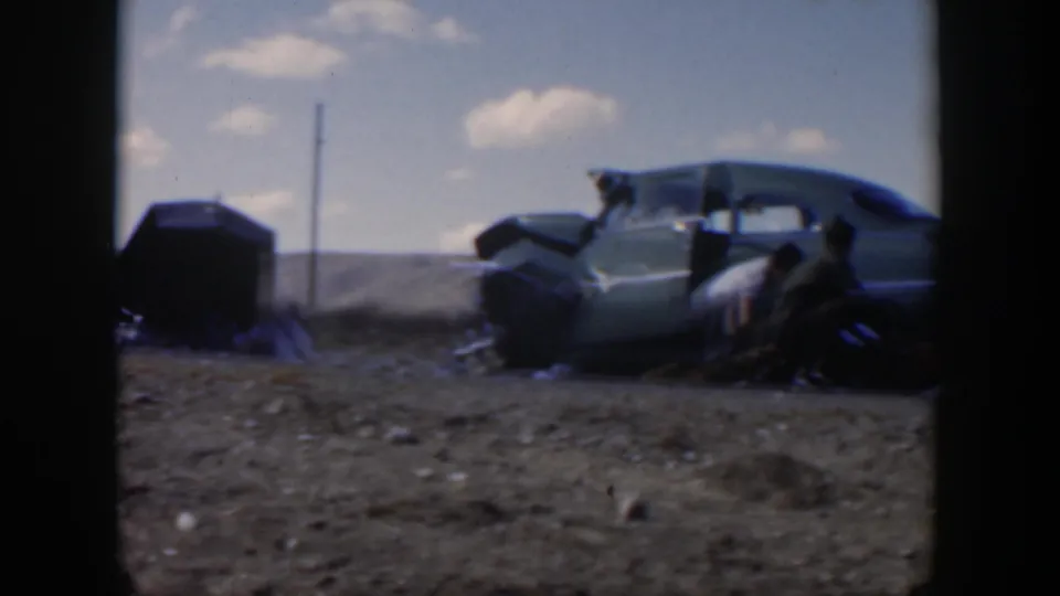 1951 Texas 8mm frame of a green sedan stopped beside a dusty road, with men positioned around the car during roadside work.