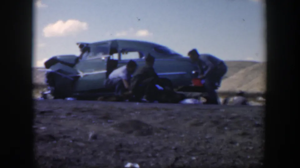 1951 Texas 8mm frame showing men working beside a green sedan on a rural roadside, with tools or bags on the ground.