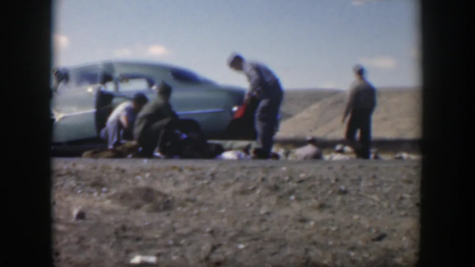 1951 Texas 8mm frame with four men around a green sedan during a roadside tire change in an open rural setting.