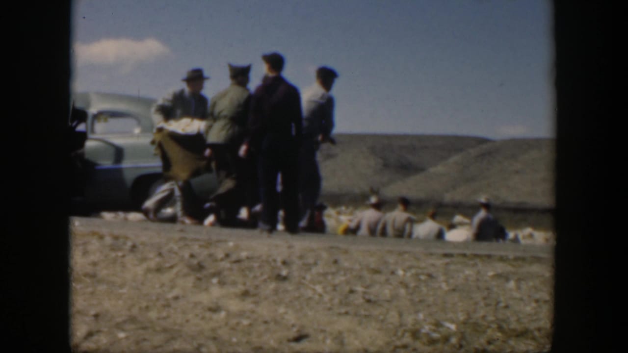 Men gather in open country as a person is carried near a roadside car.