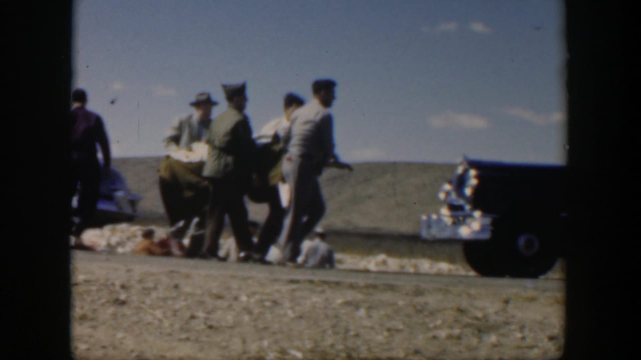 A group of men carries a person across a roadside area toward a dark vehicle.