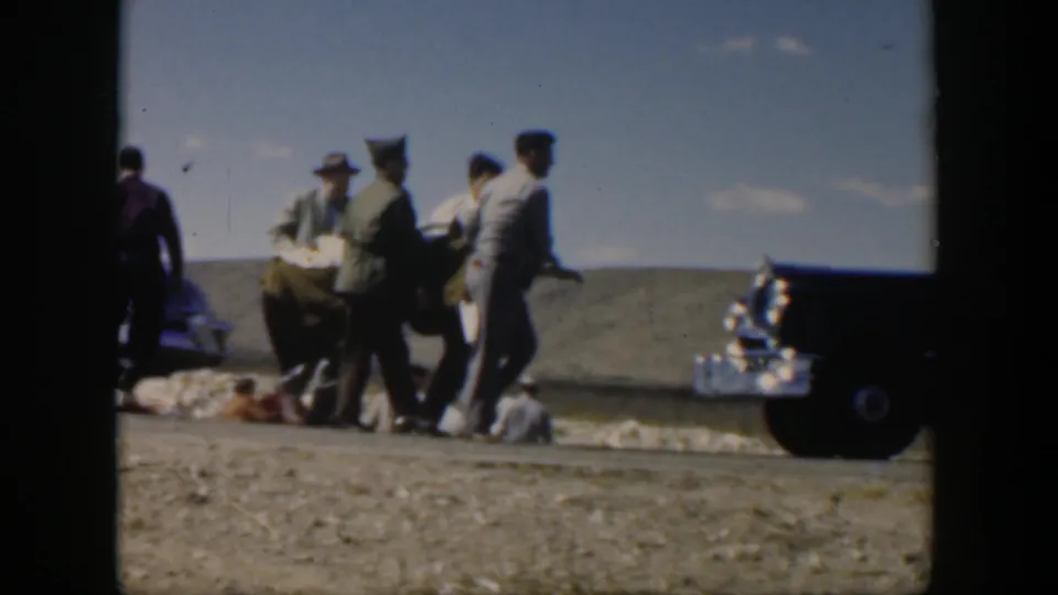Several men in 1950s attire appear to carry a person on a stretcher in an open Texas roadside setting.