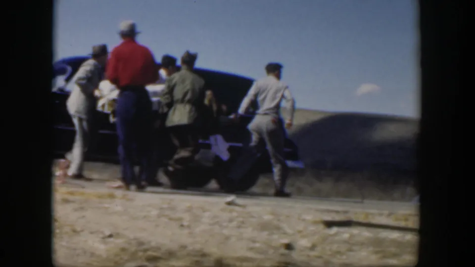 Men gather tightly at a dark car in the 1951 Texas roadside scene, appearing to load or assist a person on a stretcher.