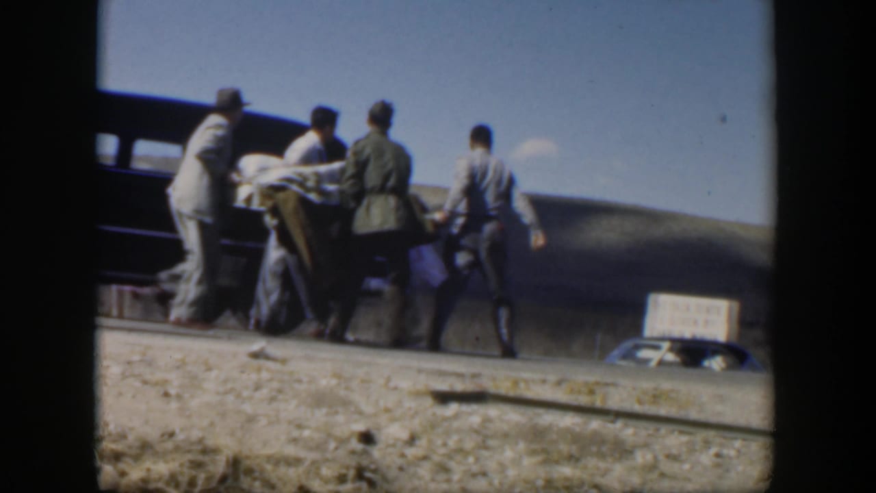 Men carry a covered stretcher toward a dark vintage sedan on a dusty roadside.