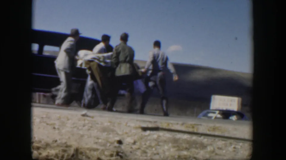 In 1951 Texas home-movie footage, several men carry a covered stretcher toward a dark sedan in a rural roadside setting.