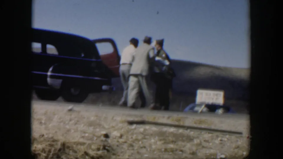 Men in period clothing gather at the open door of a dark sedan while handling a covered stretcher in rural Texas.