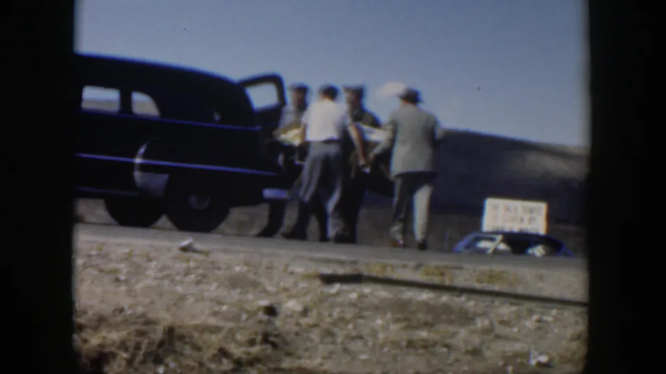 Several men cluster beside a dark vintage sedan, partly obscuring a stretcher during the roadside transfer.