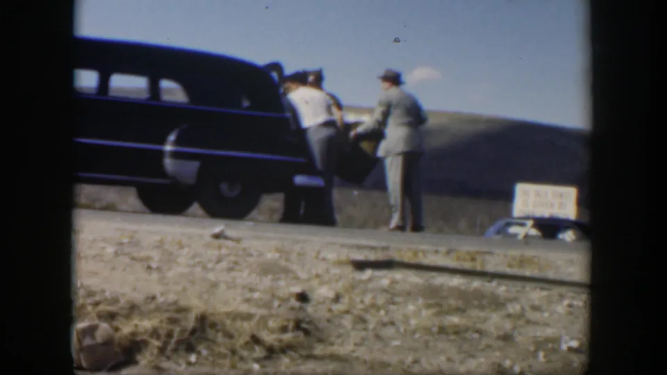 Men appear to lift or guide a covered stretcher at the side of a dark sedan on a dusty Texas roadside.