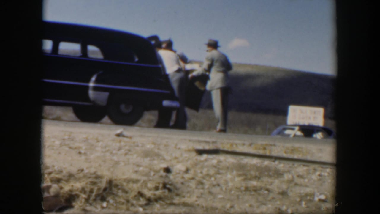 Two men stand beside a dark vehicle on a rural roadside with a small sign nearby.