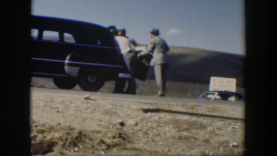 1951 Texas 8mm frame showing two men beside a dark sedan on a dry rural roadside, with open land and a small sign in the background.