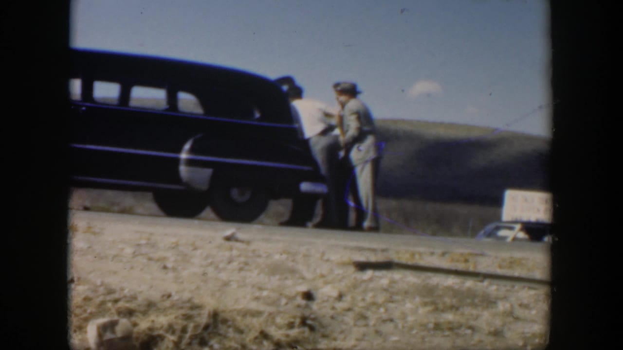 Men stand close to a dark sedan on a rural road with a small sign in the background.