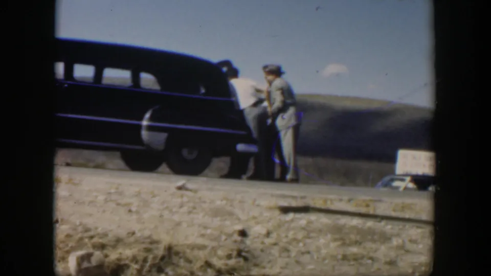 1951 Texas 8mm frame with two men near a dark sedan on a rural roadside, shot from a low bystander viewpoint across dry ground.