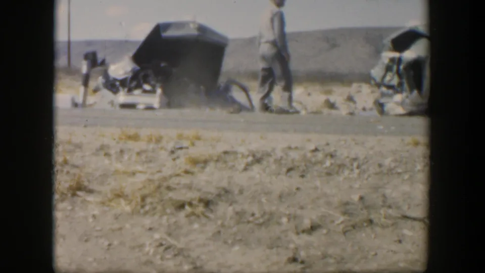1951 Texas 8mm frame showing an overturned dark car with crushed bodywork, debris on the road shoulder, and men walking or bending near the wreckage.